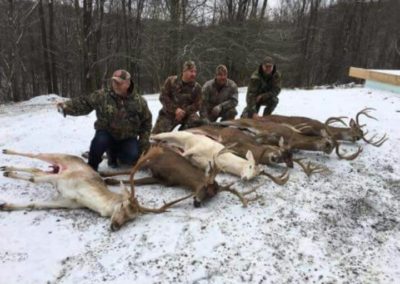 6 bucks on snowy ground with 4 kneeling hunters